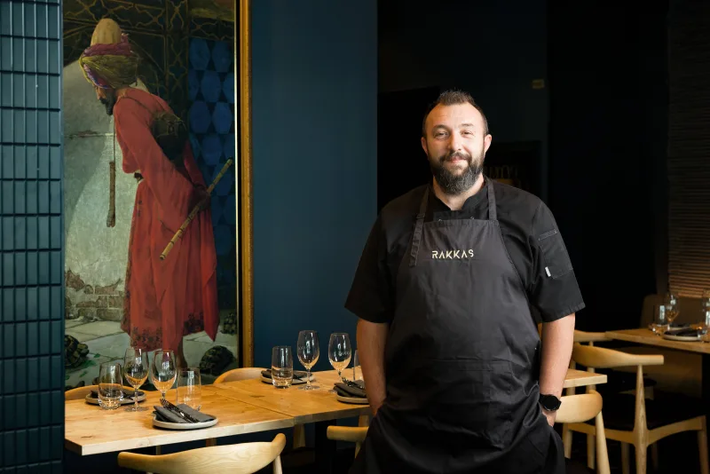 Chef Mert Toplu smiling in his RAKKAS apron, standing by the set tables with the painting on the blue wall behind him